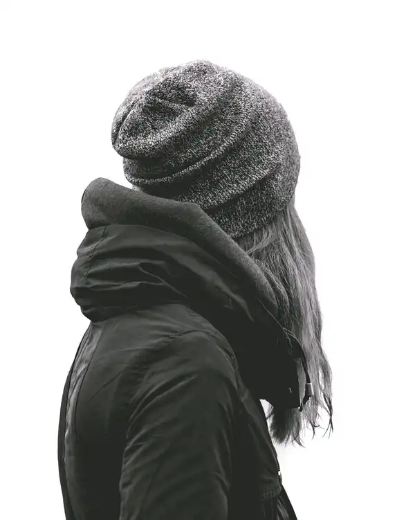 A woman in a grey marl knit beanie and a black hooded coat against a white wall