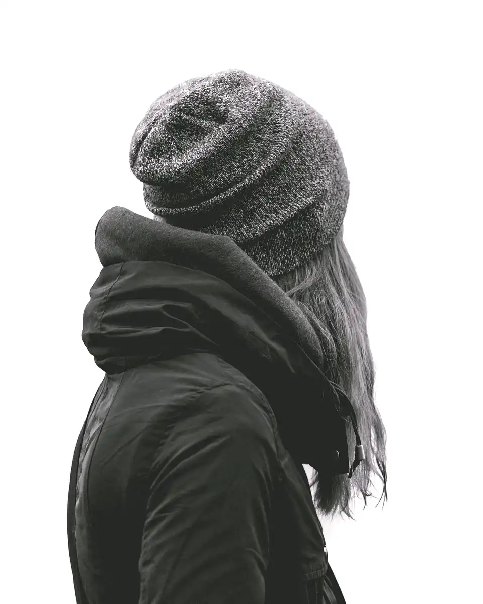 A woman in a grey marl knit beanie and a black hooded coat against a white wall