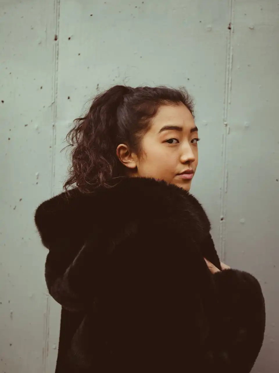 A woman in a black faux-fur coat looking back over her shoulder against a concrete wall