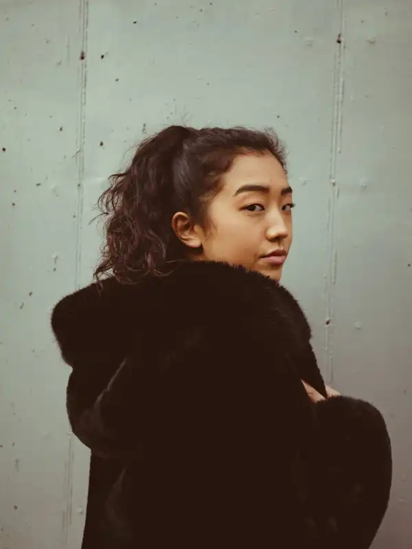 A woman in a black faux-fur coat looking back over her shoulder against a concrete wall
