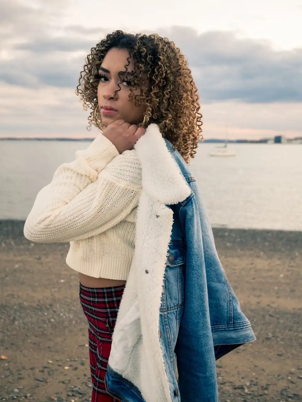 A woman in a cream chunky knit and a fleece-lined denim jacket on a grey beach