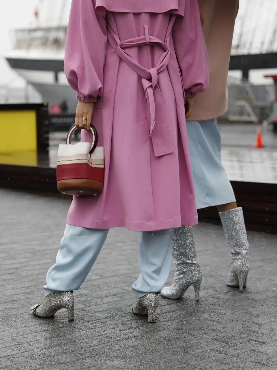 A woman in a rose pink belted coat, pale trousers and silver boots on a wet harbourside