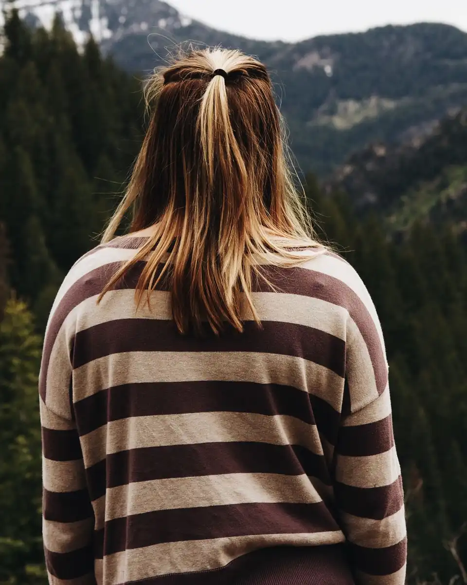A woman seen from behind in a brown and cream striped sweater, looking over a forested valley