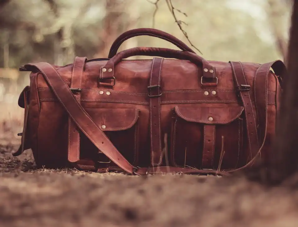 A brown leather weekend bag with buckled straps and a shoulder strap on the ground under a tree